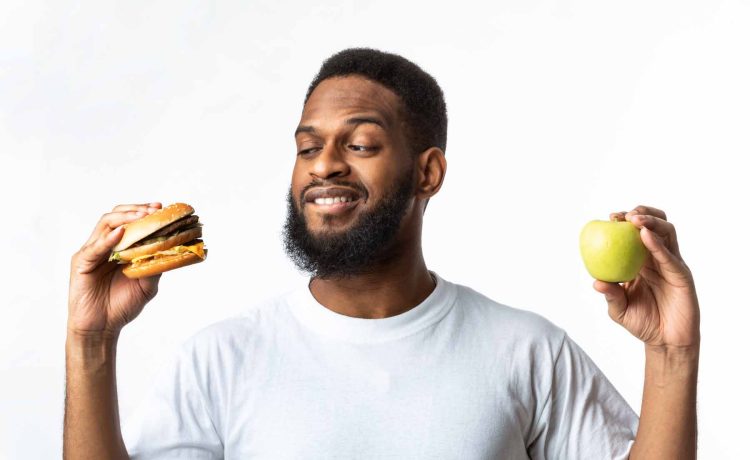 Healthy Vs Unhealthy Food. Hungry African Man Choosing Between Burger And Apple Standing In Studio Over White Background. Cheat Meal On A Diet, Male Nutrition And Junk Food Concept