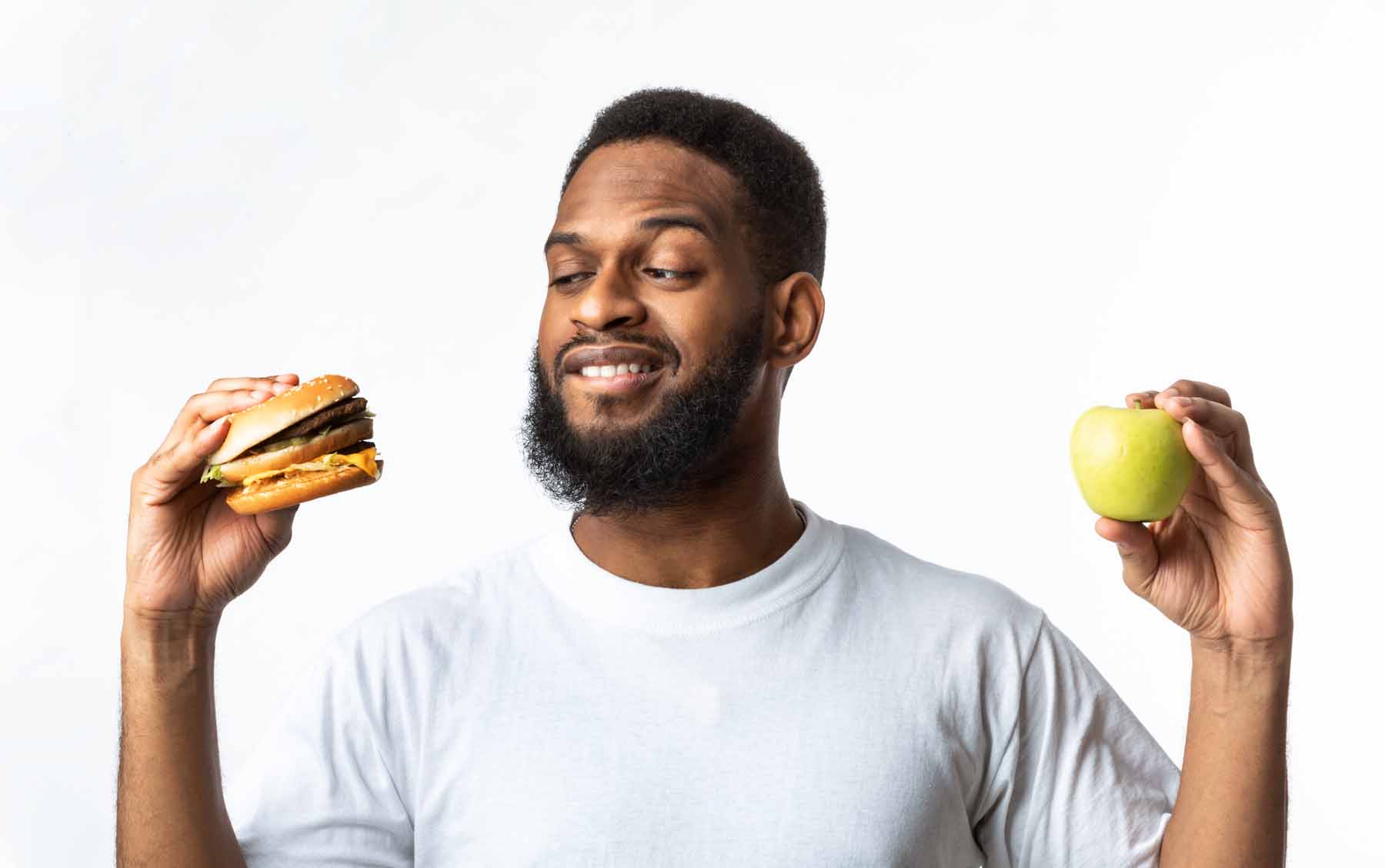Healthy Vs Unhealthy Food. Hungry African Man Choosing Between Burger And Apple Standing In Studio Over White Background. Cheat Meal On A Diet, Male Nutrition And Junk Food Concept